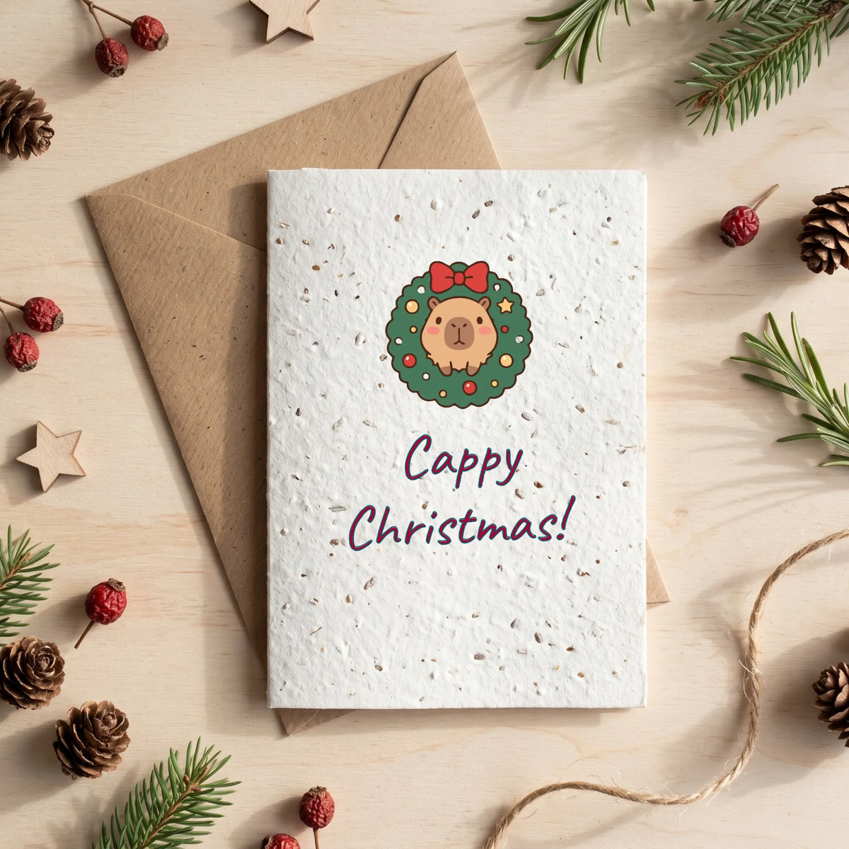 A square white Christmas greeting card with a textured speckled finish, featuring a cartoon capybara inside a green wreath with a red bow and the words 'Cappy Christmas!'. The card is displayed on top of a brown kraft envelope, surrounded by pine branches, pinecones, and wooden stars on a light wood background.