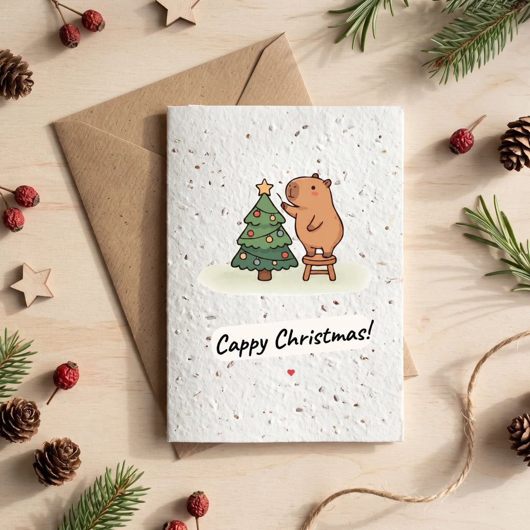 A square white greeting card with a textured paper finish lies on top of a kraft envelope, styled in a flat lay with pine branches, pinecones, and small red berries on a light wood surface. The card features a cartoon capybara standing on a stool and painting a Christmas tree, with the text &