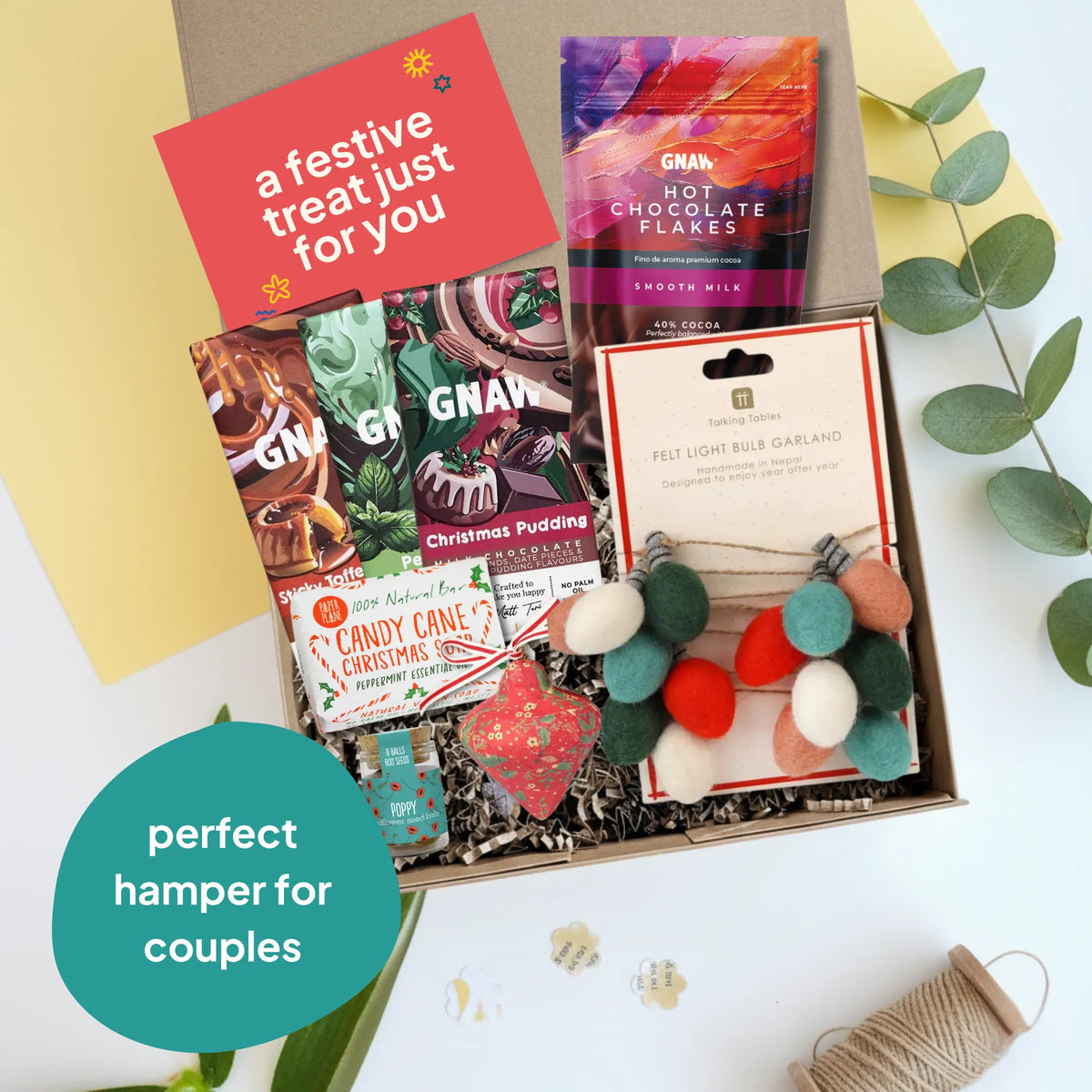 A curated gift hamper box for couples, filled with hot chocolate flakes, Christmas pudding chocolate, a felt garland, a red felt heart decoration, arranged in a cardboard box with shredded paper. The box is styled on a white surface with a yellow background panel, eucalyptus leaves, and a spool of twine.