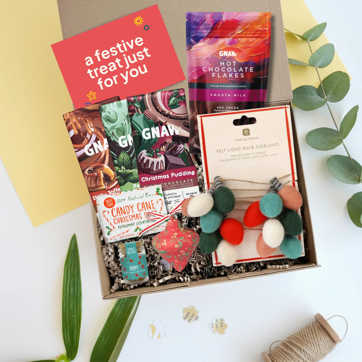 A curated gift box filled with festive treats and decorations, photographed from above on a white surface with decorative leaves and paper. The box contains a GNAH hot chocolate package, three Christmas themed chocolate bars , a candy cane Christmas soap, a felt light bulb garland, a red heart ornament, and a red greeting card that reads 'a festive treat just for you'.