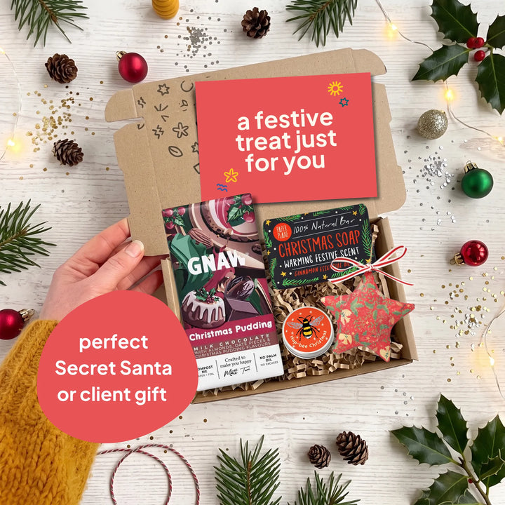 A hand holds open a kraft gift box filled with holiday treats on a white wood surface decorated with pine branches, ornaments, and fairy lights. The box contains a red card reading &
