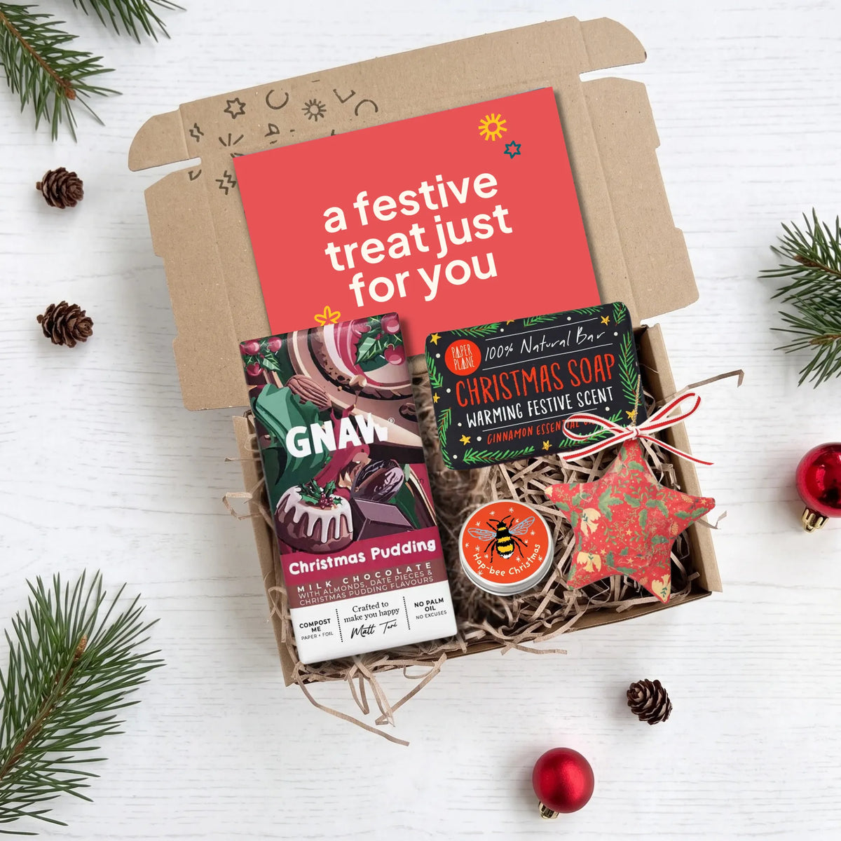 A Christmas-themed gift box filled with treats on a white wood background. The open kraft cardboard box contains a red card that reads 'a festive treat just for you,' a packaged chocolate bar labeled 'GNAW,' a tin of '100% Natural Bar' Christmas soap in 'Cinnamon Lime,' and a red star-shaped ornament tied with a red ribbon.