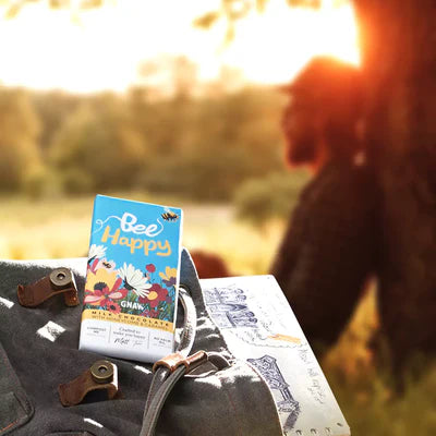 'Bee Happy' Honeycomb + Caramel Milk Chocolate Bar in colourful packaging with illustrations of flowers and bees. In the background a man sits under a tree in a meadow. 