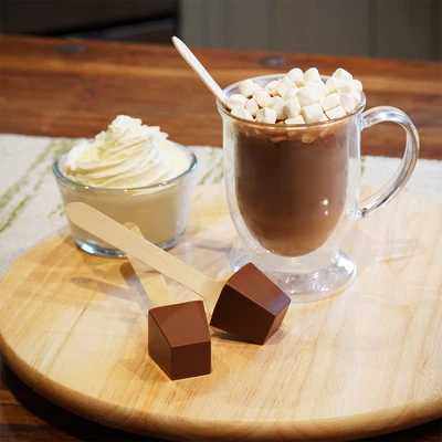 Hot chocolate with marshmallows in a clear mug on a wooden table, surrounded by whipped cream and chocolate pieces.
