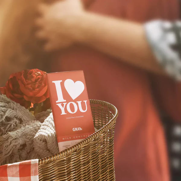 ethical milk chocolate bar by Gnaw in a dark red wrapper with white I Love You illustration. The chocolate is in a gift basket with a red rose. 