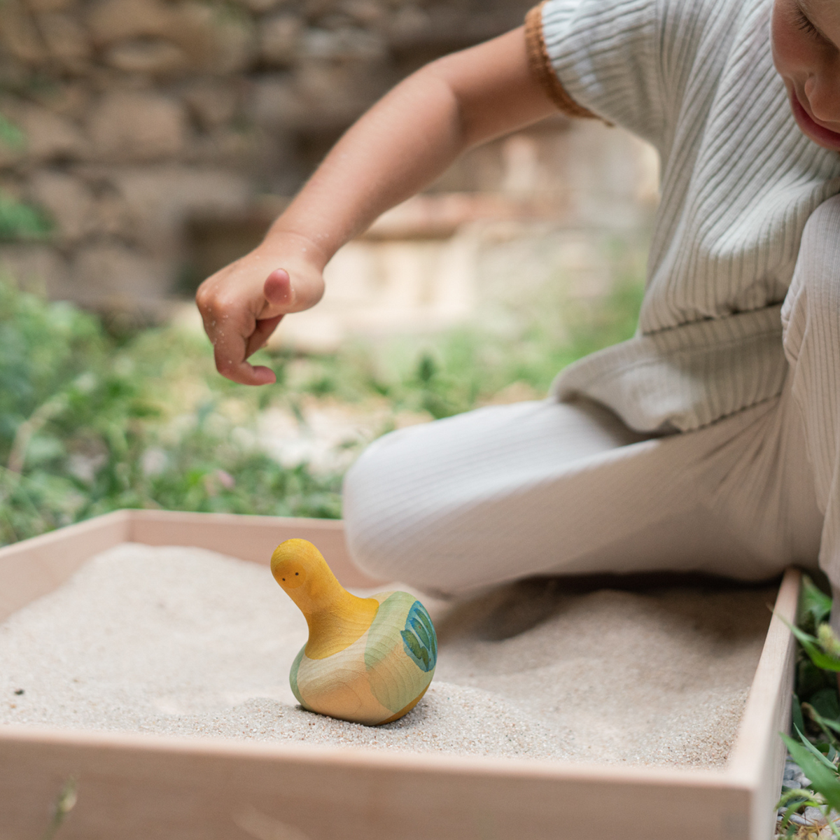 child playing with a yellow bird-themed wooden spinning top toy in a play tray filled with sand. The toys are Made in spain by Grapat toys
