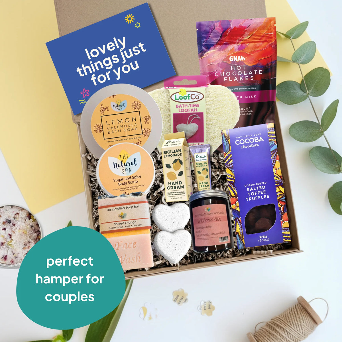 A curated gift hamper box for couples, filled with various wellness and snack items including a blue card reading 'lovely things just for you', LooFoo Lotion, The Natural Spa body wash, Nic & Julian Cream, a jar of chocolate truffles, and a box of cocoa chocolate. The box is styled on a white surface with decorative leaves, twine, and a teal badge that says 'perfect hamper for couples'.