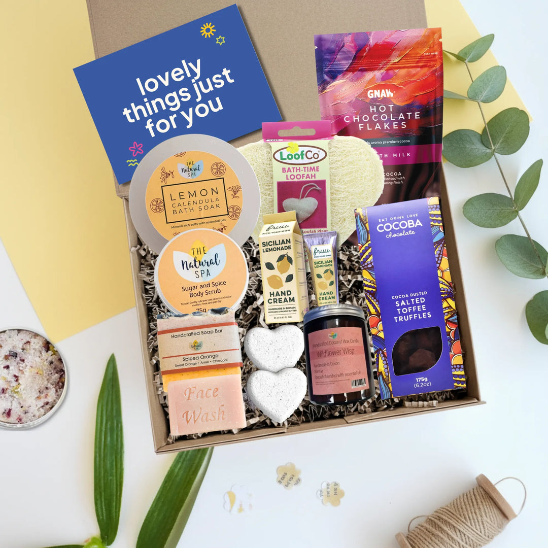A curated gift box filled with assorted self-care and snack items, photographed from above on a white surface with decorative leaves and paper. The box contains a blue card reading &