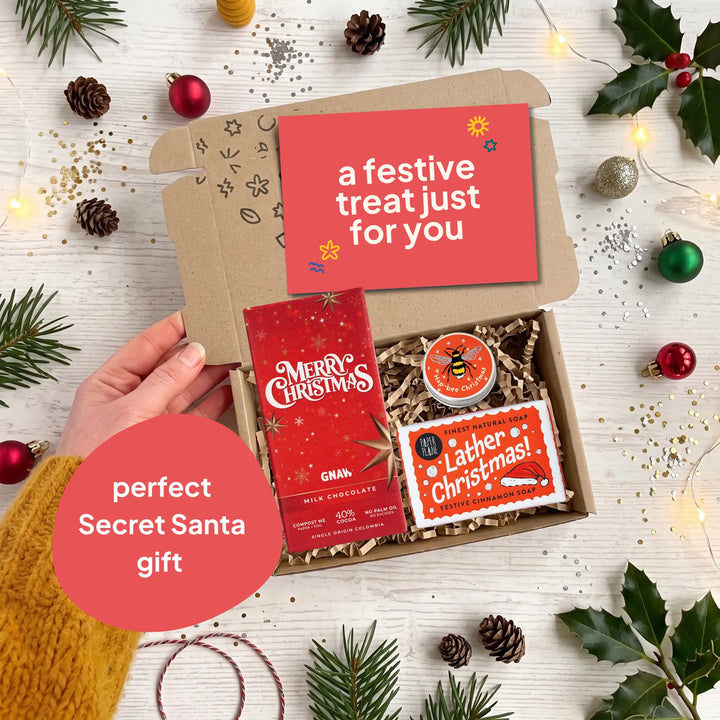 A Christmas-themed gift box opened by a hand, filled with festive treats including a red &