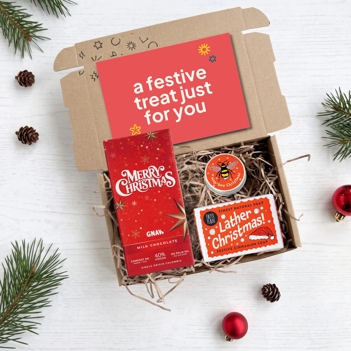 A curated Christmas gift box filled with treats on a white wood background, surrounded by pine branches and ornaments. The open kraft box contains a red chocolate bar labeled 'Merry Christmas', a small round tin with a bee logo, and a cinnamon soap bar with 'Lather Christmas!' packaging.