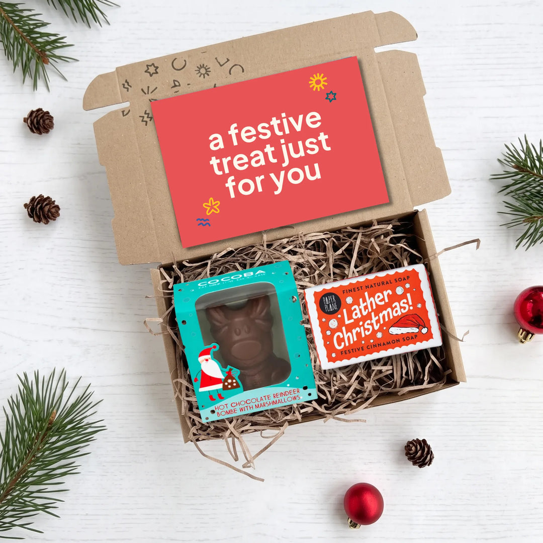 An open kraft gift box filled with shredded paper on a white wood surface, surrounded by pine branches, pinecones, and red ornaments. Inside the box is a hot chocolate bomb, a packaged dark Christmas soap bar and a small red holiday card that reads &