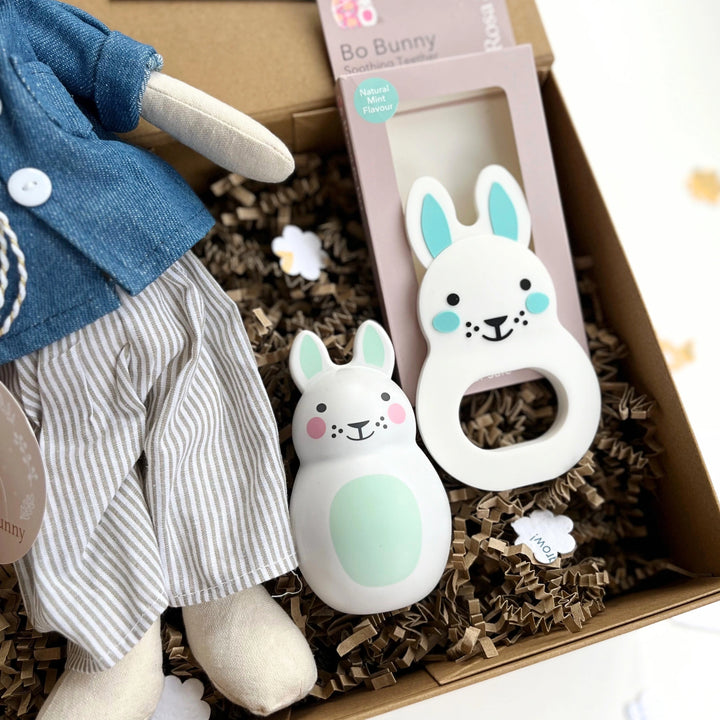 Baby toys including a plush toy, two rabbit-shaped rattles, and a gift box with brown tissue paper.