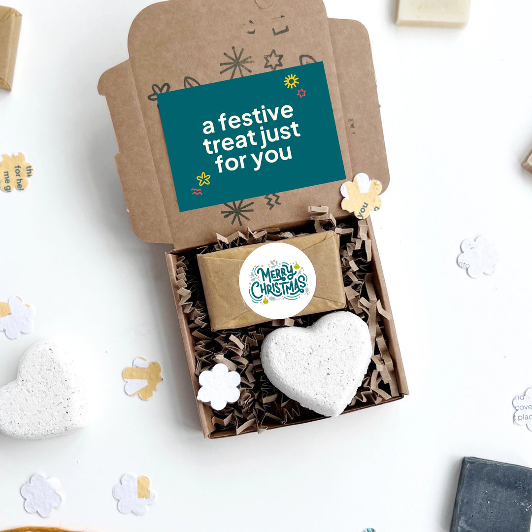 Gift box with a heart-shaped bath bomb, mini soap bar with 'merry christmas' message and a small box, on a white background with decorative elements.