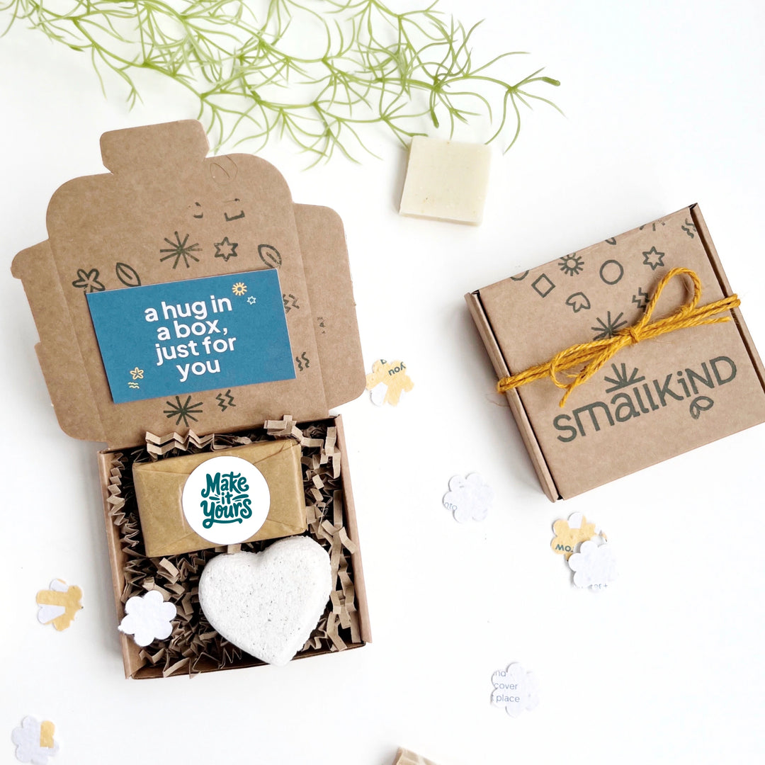 SmallKind gift box with heart-shaped bath bomb, branded mini soap and 'a hug in a box, just for you' text on a card, all on a white background.