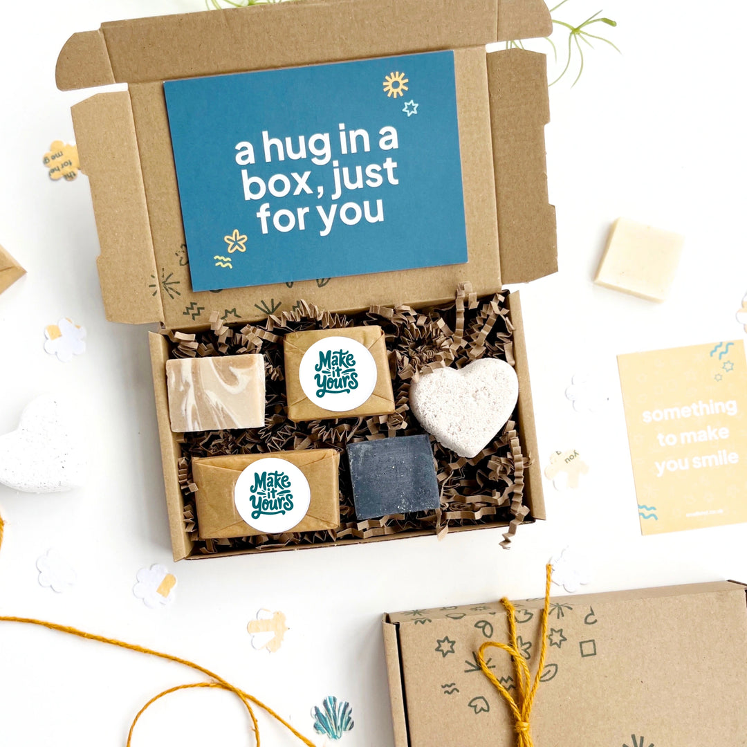 Gift box with self-care products including soap and a heart-shaped bath bomb, with a card inside reading 'a hug in a box, just for you'.