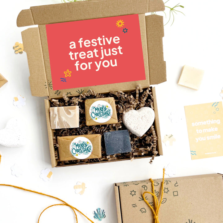 Gift box with festive self care items including a christmas soap and a heart-shaped bath bomb on a white background.