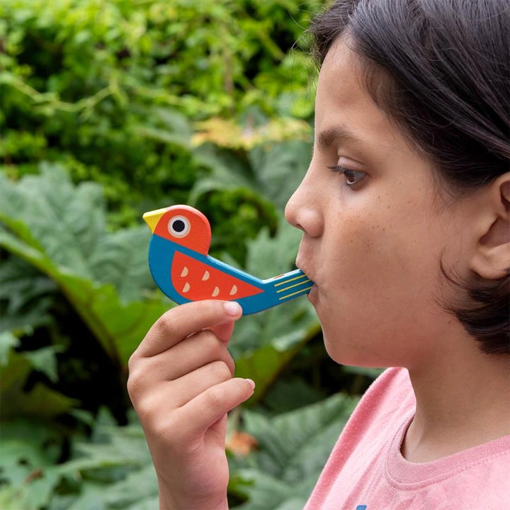 girl with white skin and dark hair blowing a Rex London colourful wooden bird shaped whistle in red/blue. 