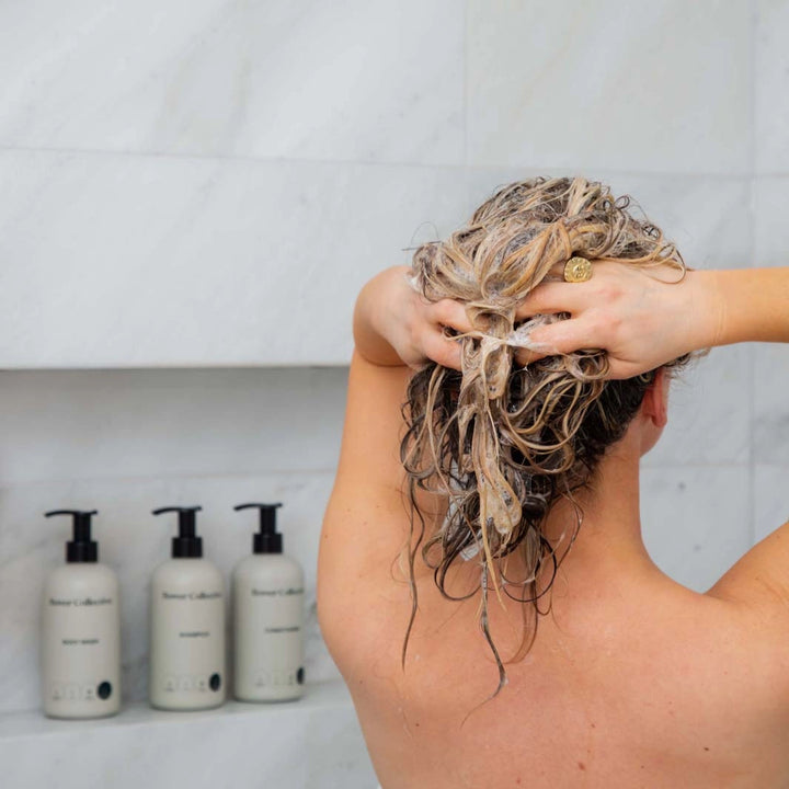 woman with long blond hair washing her hair with bottle of bower collective shampoo and conditioner in the background