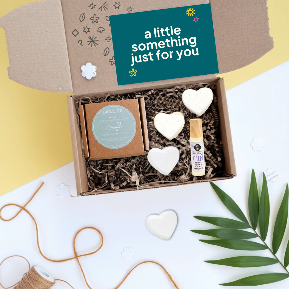An open gift box on a white surface with a yellow corner backdrop, containing a Calm essential oil roller, pack of 4 coconut wax melts and three white heart-shaped bath bombs. The inner lid has a teal card reading 'a little something just for you' with doodles.