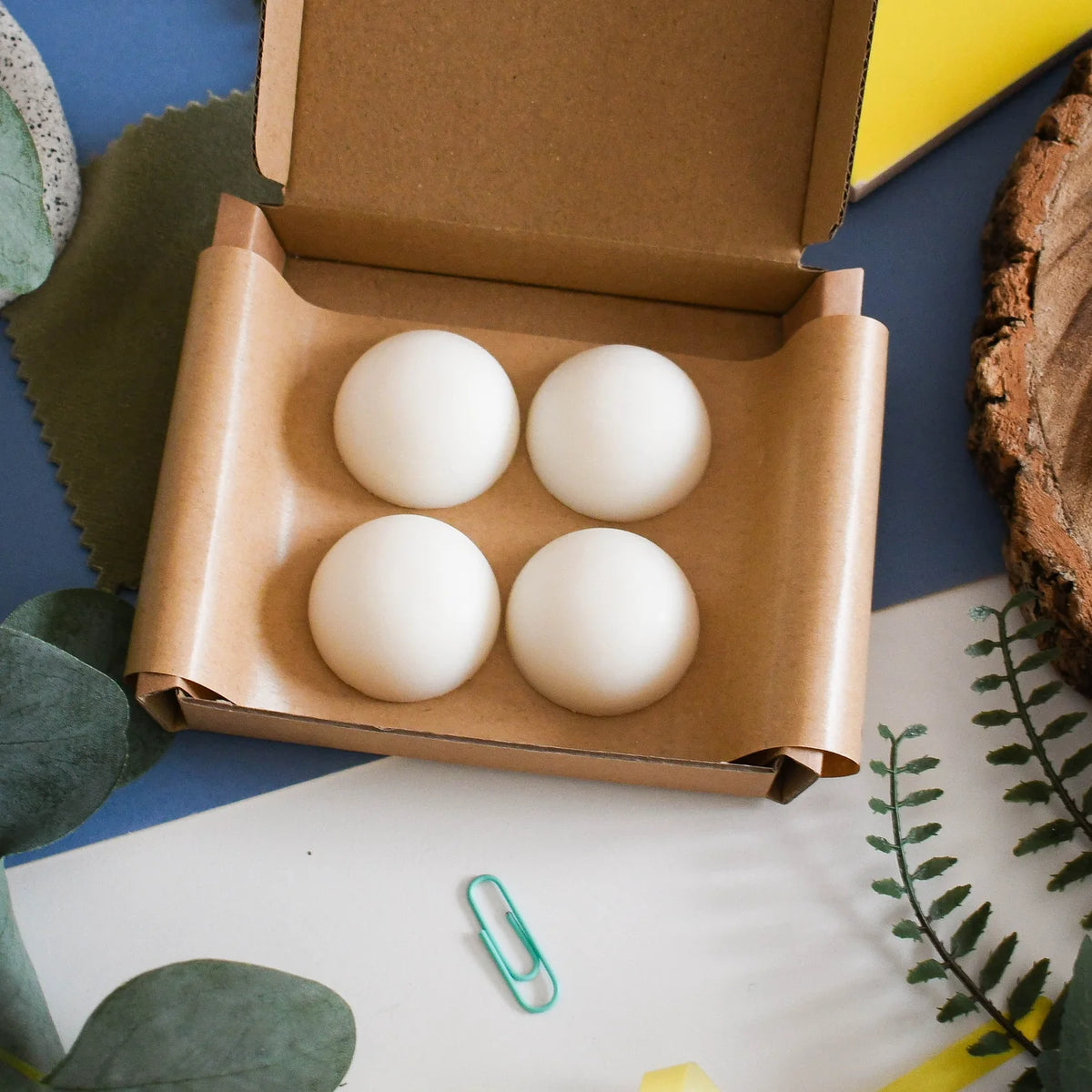 Four smooth white wax melts arranged inside an open brown cardboard box lined with brown kraft paper, photographed from above on a styled flat-lay surface with leaves, paper clips, and wood accents.