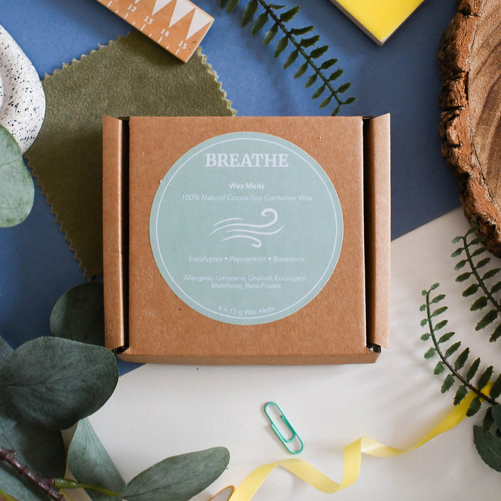 A square kraft paper box labeled BREATHE Wax Melt, styled in a flat-lay arrangement with green leaves, a yellow ribbon, a green paperclip, textured paper, and wood on a blue and white background.