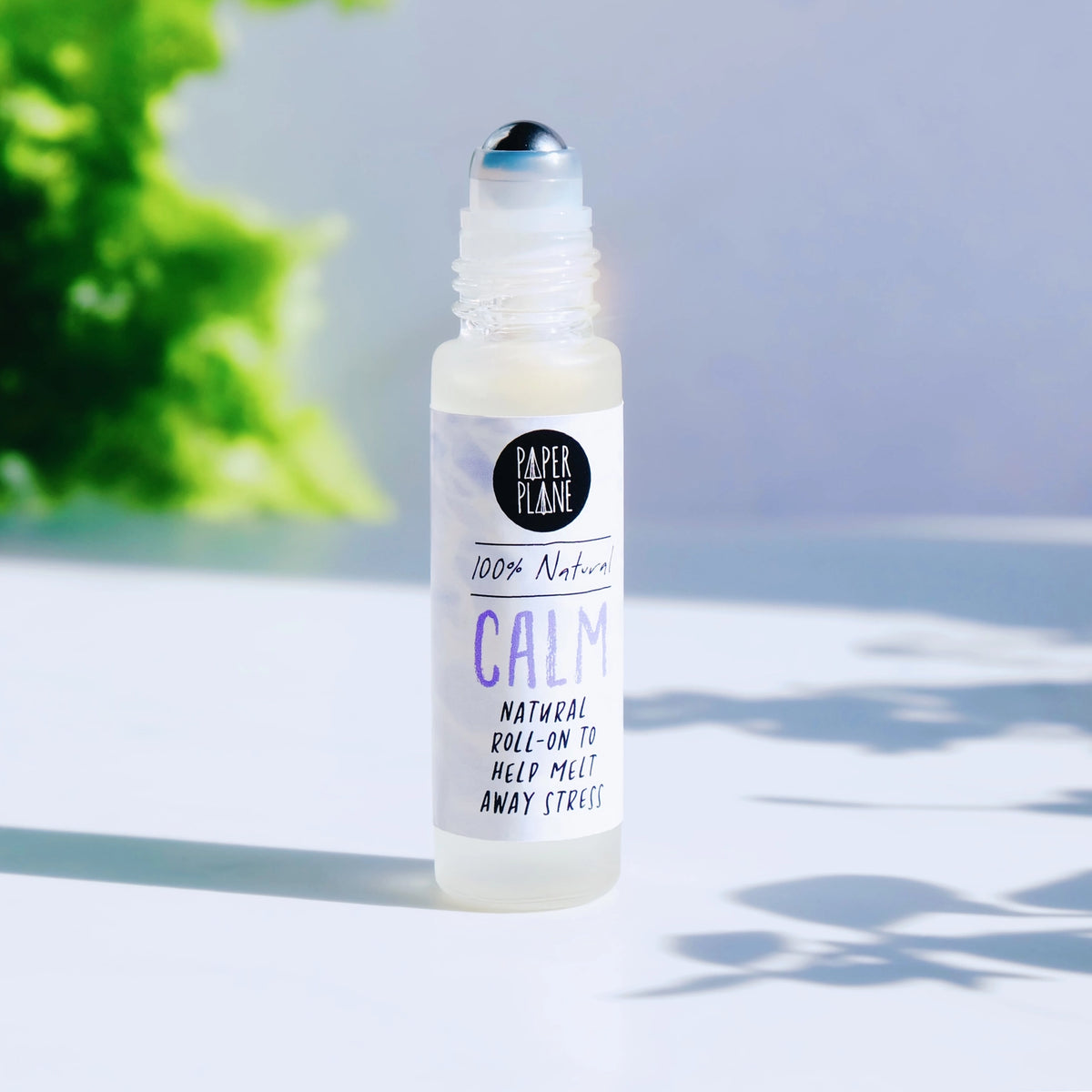 A clear glass roll-on bottle with a white label reading 'Paper Plane' and 'Calm' in lavender and black text, photographed on a bright white surface with soft green foliage in the background.