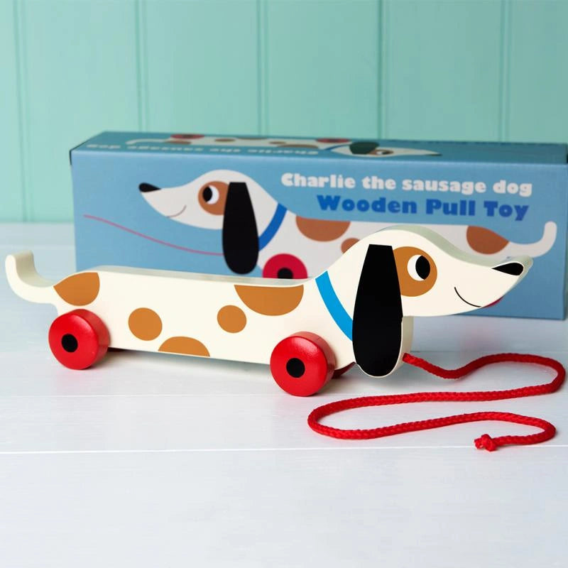 brown and white painted wooden sausage dog pull along toy with red wheels and a red string. The toy is in front of a blue retro style gift box. For children over 3 years