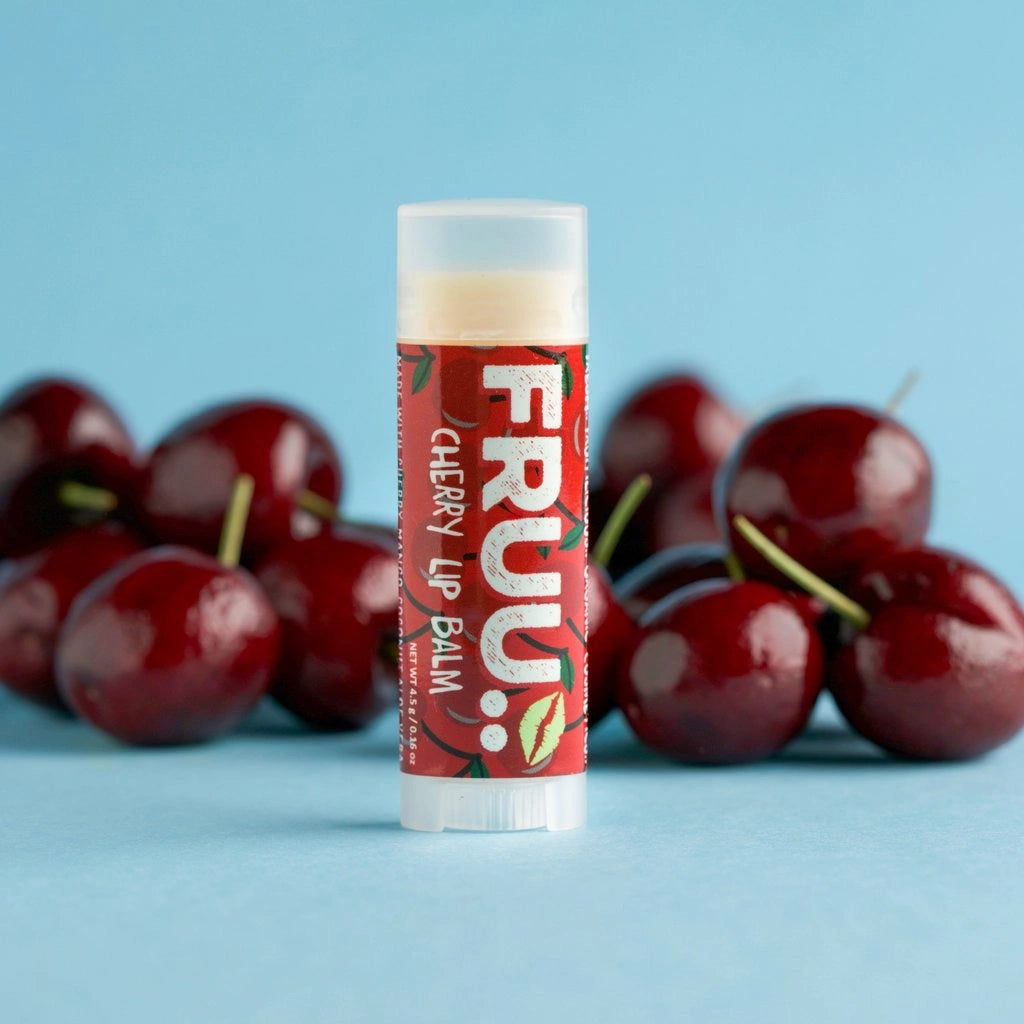 A tube of FRUU Cherry Lip Balm stands upright in the center, with glossy red cherries blurred in the background against a light blue backdrop.