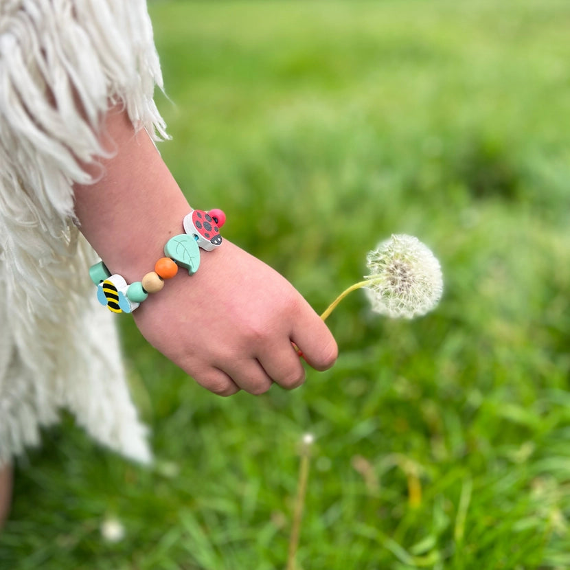 Child's hand holding a dandelion with a colourful minibeast beaded bracelet on a grassy background