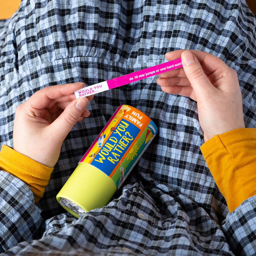 Person holding a 'Would You Rather?' question stick and container, with a checkered shirt in the background.