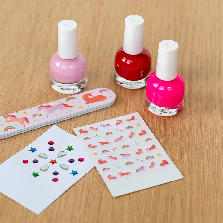 A nail art kit on a light wood surface, including three nail polish bottles in pink, red, and bright pink, a unicorn-themed nail file, and three sheets of decorative stickers with stars, rainbows, and unicorns.