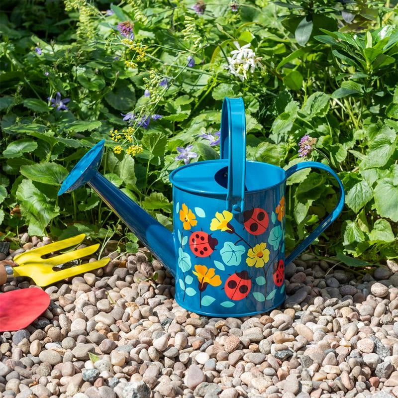 blue metal watering can with ladybird print on a gravel path in front of a flower bed. Made by rex london for kids age 3 and over. perfect gift for gardeners