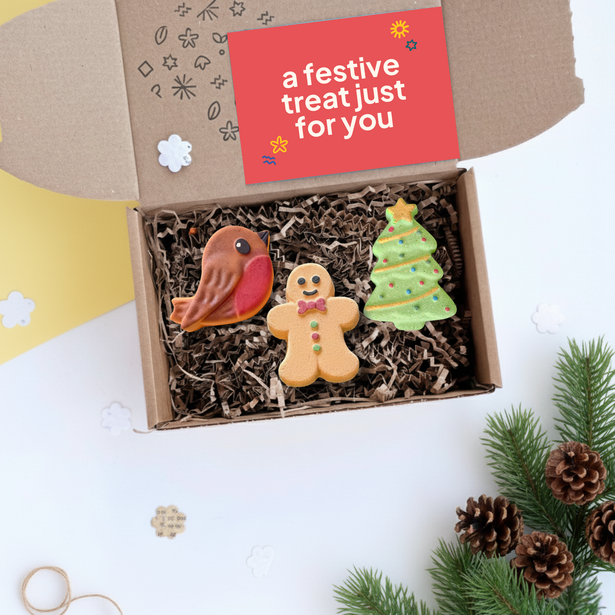 An open brown gift box filled with shredded paper and Christmas themed bath bombs, including a robin, gingerbread man, and Christmas tree, with a red card reading 'a festive treat just for you' inside. The box is styled on a white surface with pine branches and pinecones around it.