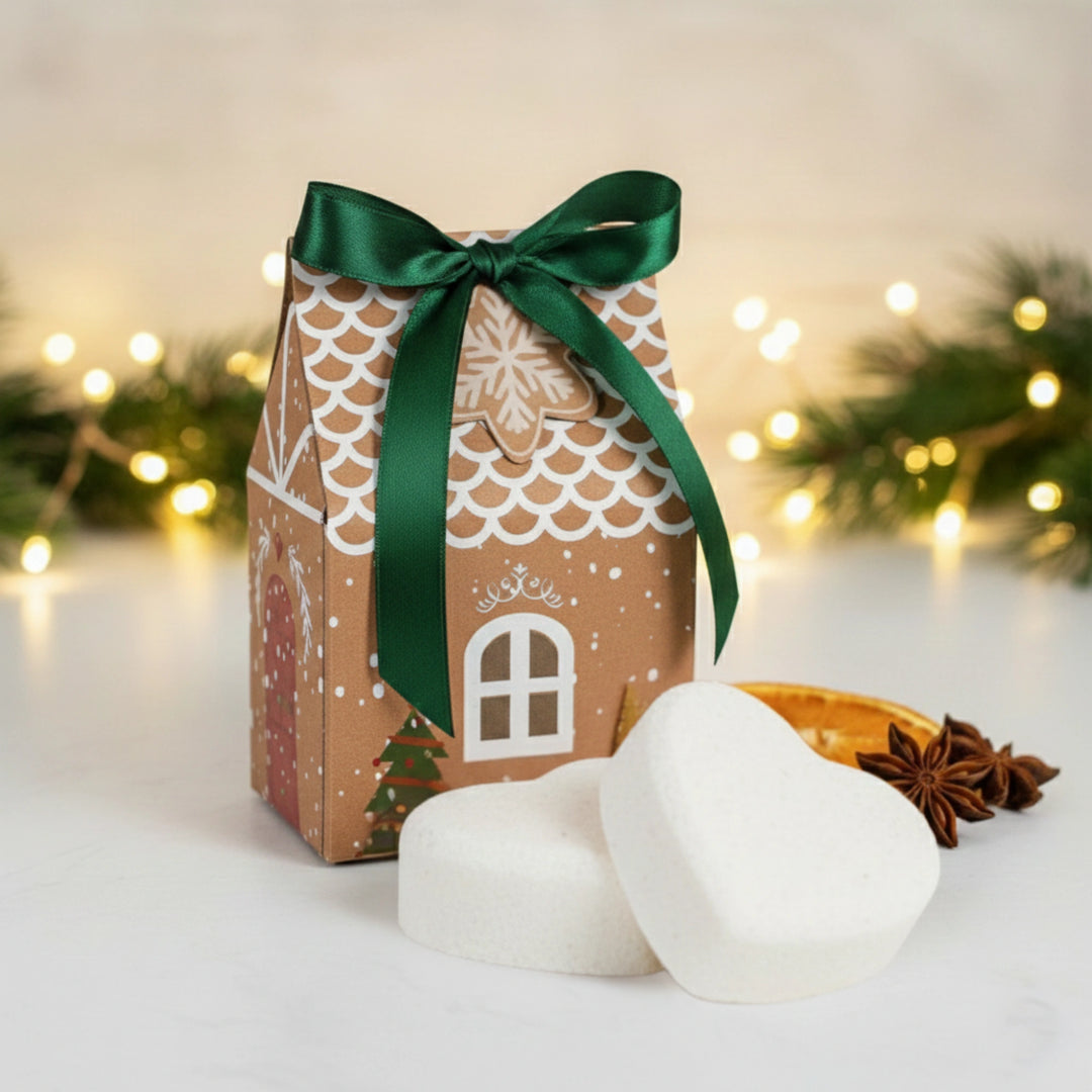 Gingerbread house-shaped gift box with a green ribbon and two heart shaped bath bombs on a festive background.