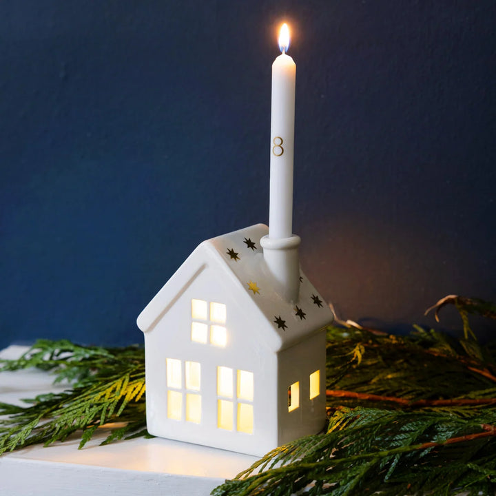 White house-shaped lantern with a lit candle on top, surrounded by greenery against a dark sky.