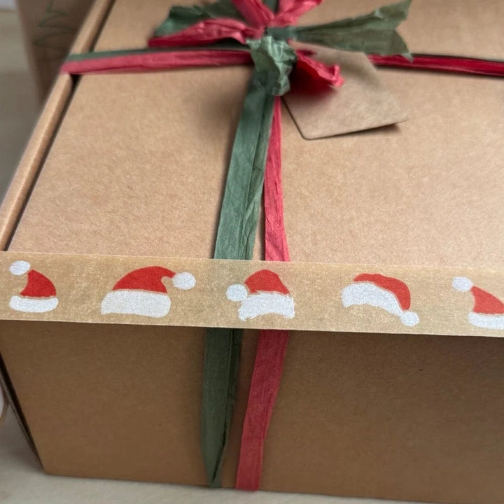Gift box wrapped in brown paper with a Santa hat festive paper tape, green and red ribbon and tag.