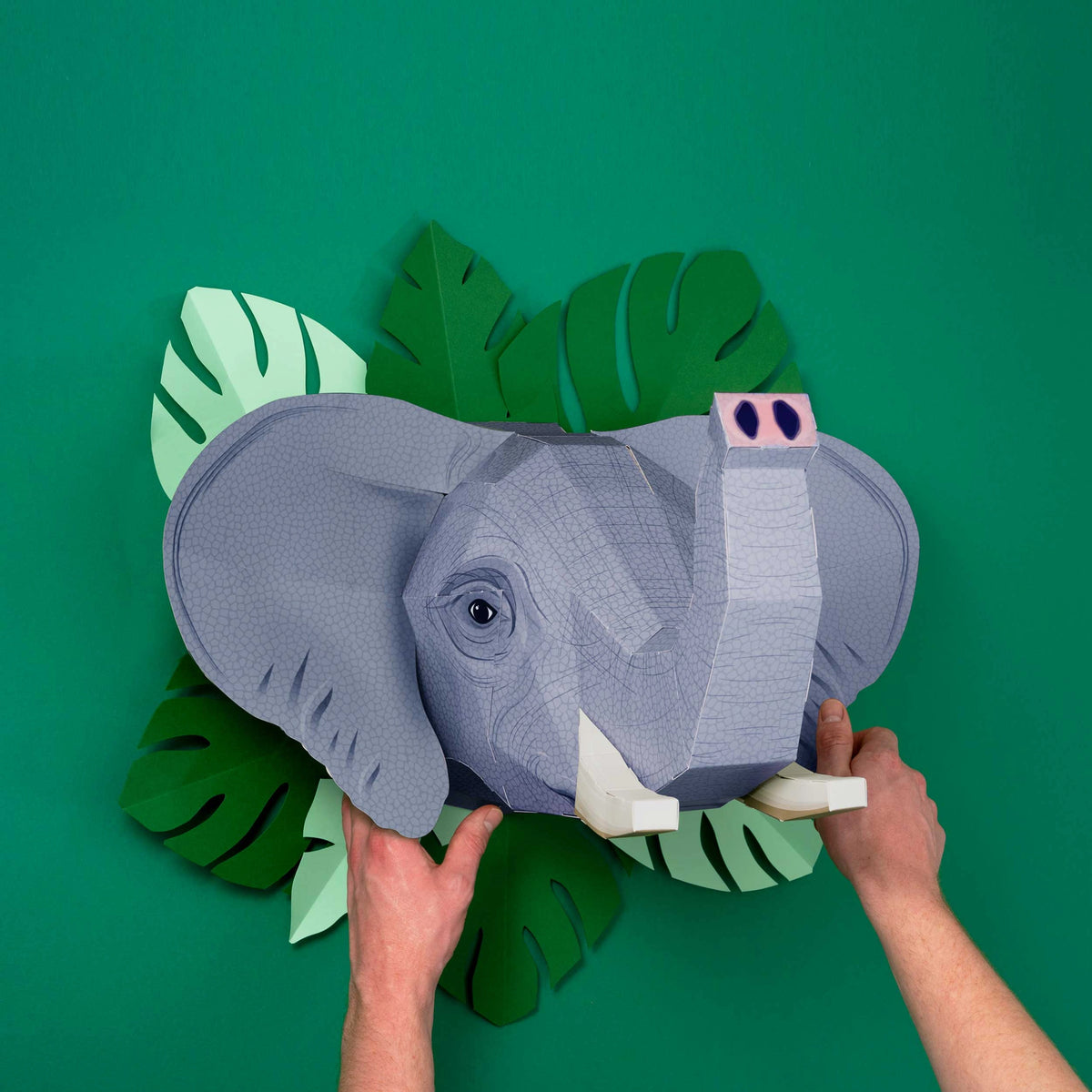front View of the Create Your Own Extraordinary Elephant Craft Kit by Clockwork Soldier. Two hands holding up the finished elephant head  in front of a green wall.