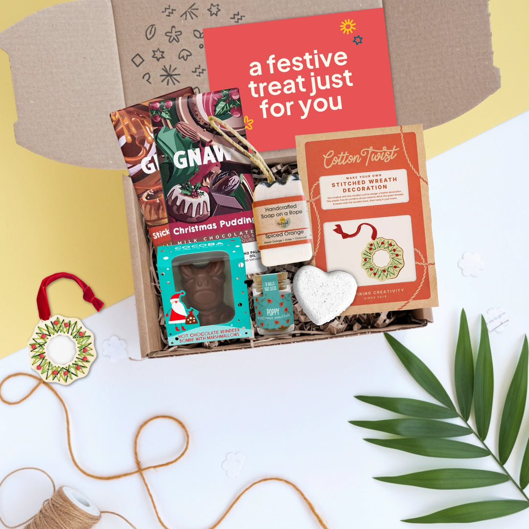 Gift box with festive items including a make your own decorations kit, chocolate bars, hot chocolate bomb, soap and seedballs on a white surface.