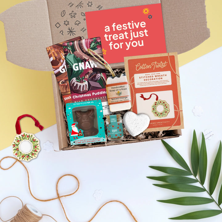 Gift box with festive items including a make your own decorations kit, chocolate bars, hot chocolate bomb, soap and seedballs on a white surface.