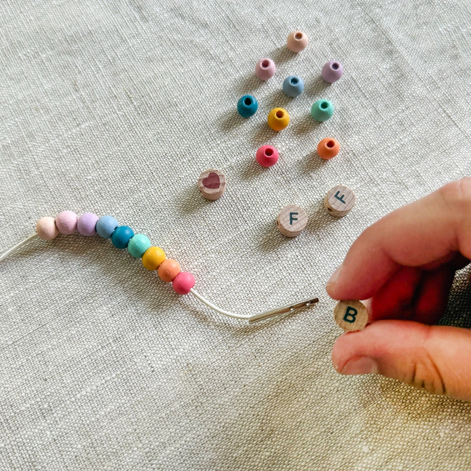 Colourful round beads and letter beads on a textured fabric surface with a hand holding a needle.
