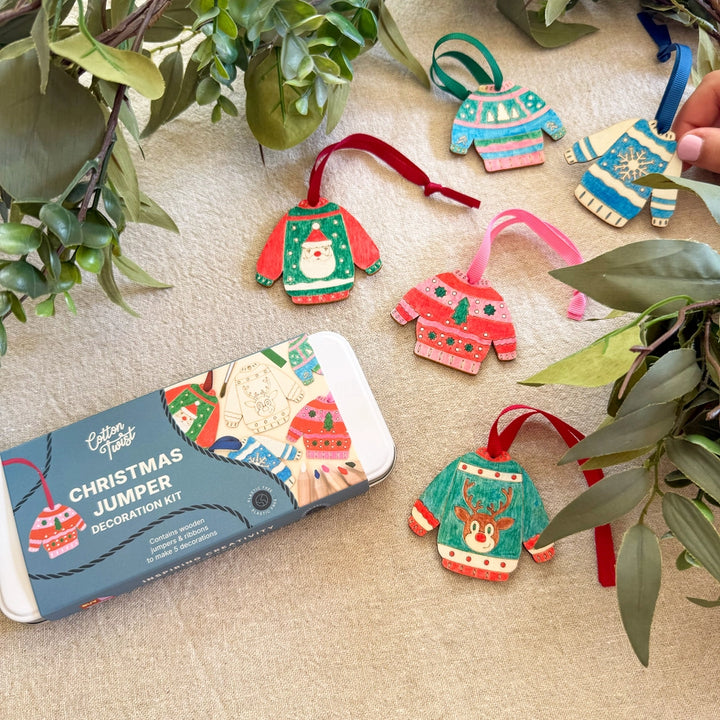 five wooden Christmas Jumper hanging decorations with a 'Christmas Jumper Decoration Kit' tin by Cotton Twist. A child's hand holds one of the decorations.