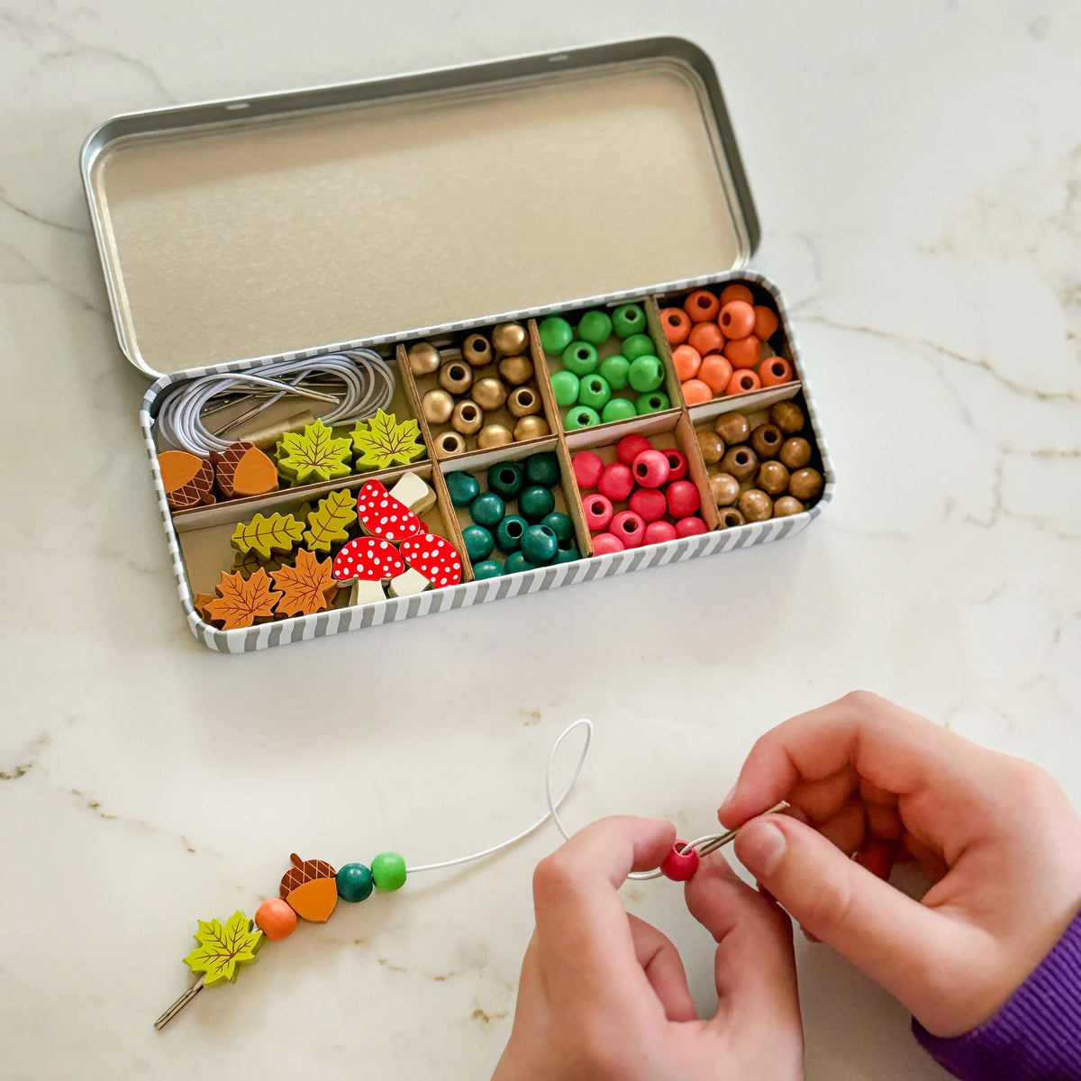 Childs hands threading leaf, acorn and round beads from the Cotton Twist Forest Bracelet Beading Kit. The gift tin sits open on a surface showcasing the nature inspired wooden beads in the kit. 