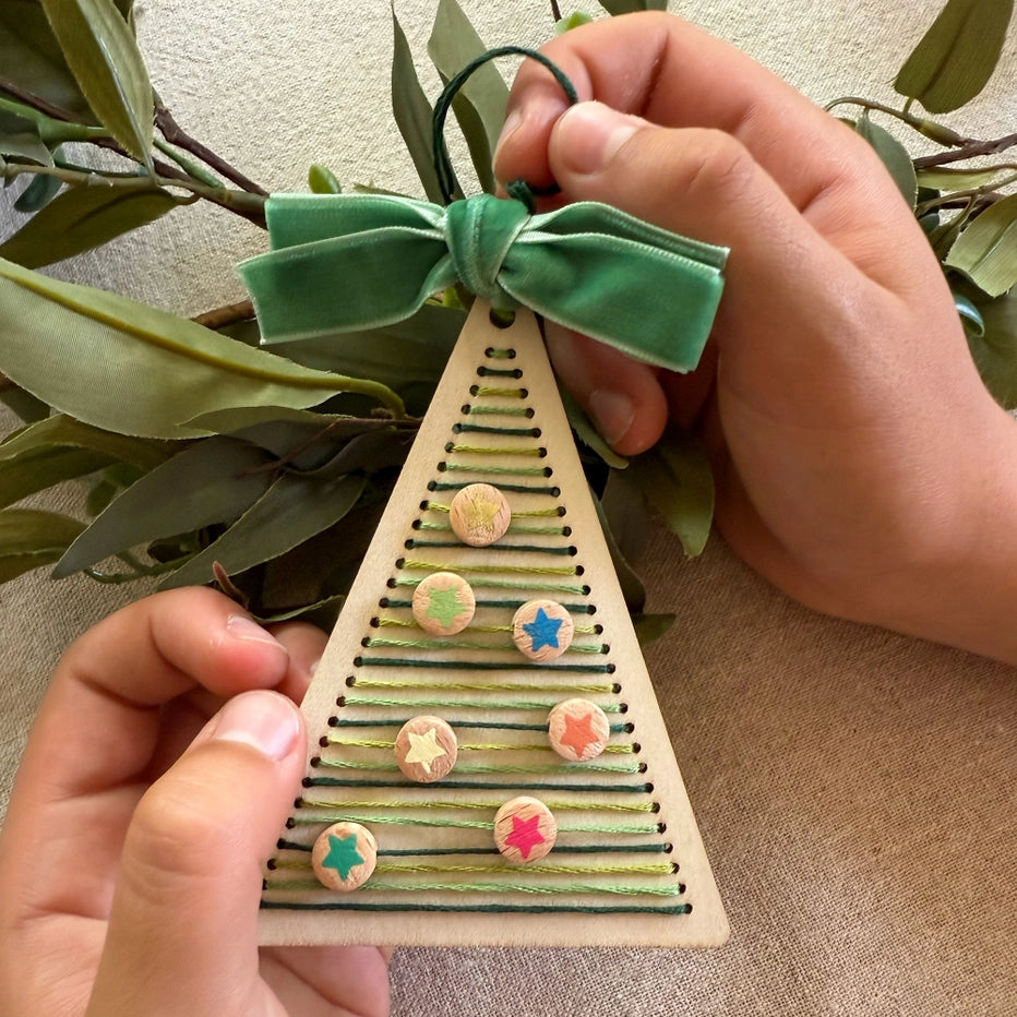 Handmade wooden Christmas tree ornament with colourful buttons and a green ribbon, held by hands against a neutral background.