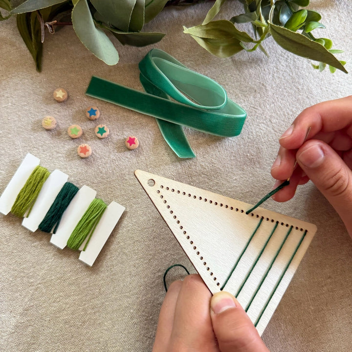 child's hands threading green thread through a wooden embroidery hoop with colourful beads and green ribbons on a  linen tablecloth