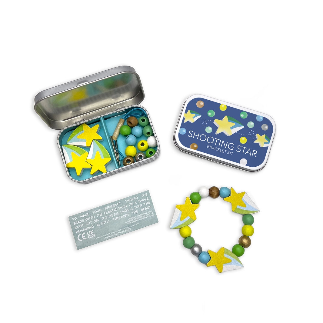 Overhead view of a Shooting Star Bracelet Kit. for children. An open rectangular tin reveals colourful wooden beads (blue, green, yellow, white), elastic with metal ends, and star-shaped beads. Next to it is the closed tin with a star and polka dot design. Below is a completed space-themed bracelet and a small instruction card.