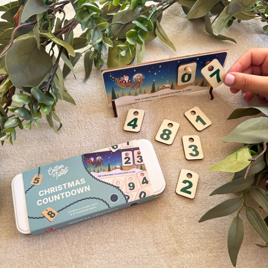 wooden Christmas countdown calendar with numbered tokens and gift tin on a table with linen tablecloth. A child's hand is placing a number 7 token on the calendar. 
