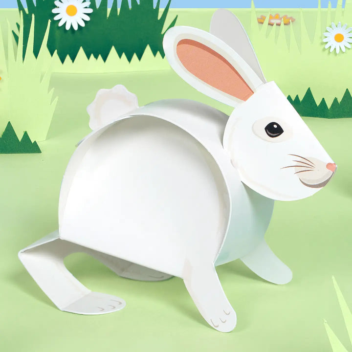A white paper cutout rabbit figure with pink inner ears, shown standing on a light green illustrated grassy background with daisies and green hills.