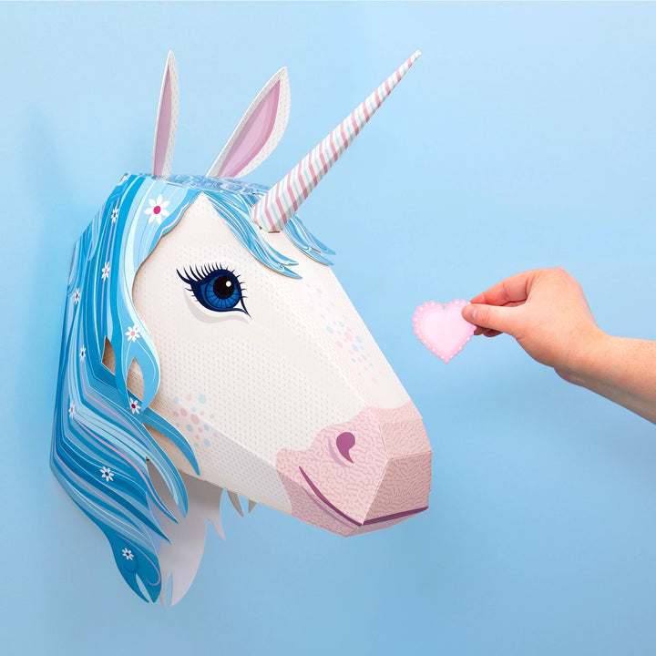 A hand holds a pink heart-shaped pendant toward a mounted paper unicorn head wall decoration. The unicorn has a white face, pink nose and horn details, blue and white hair, and pink inner ears, set against a light blue background.