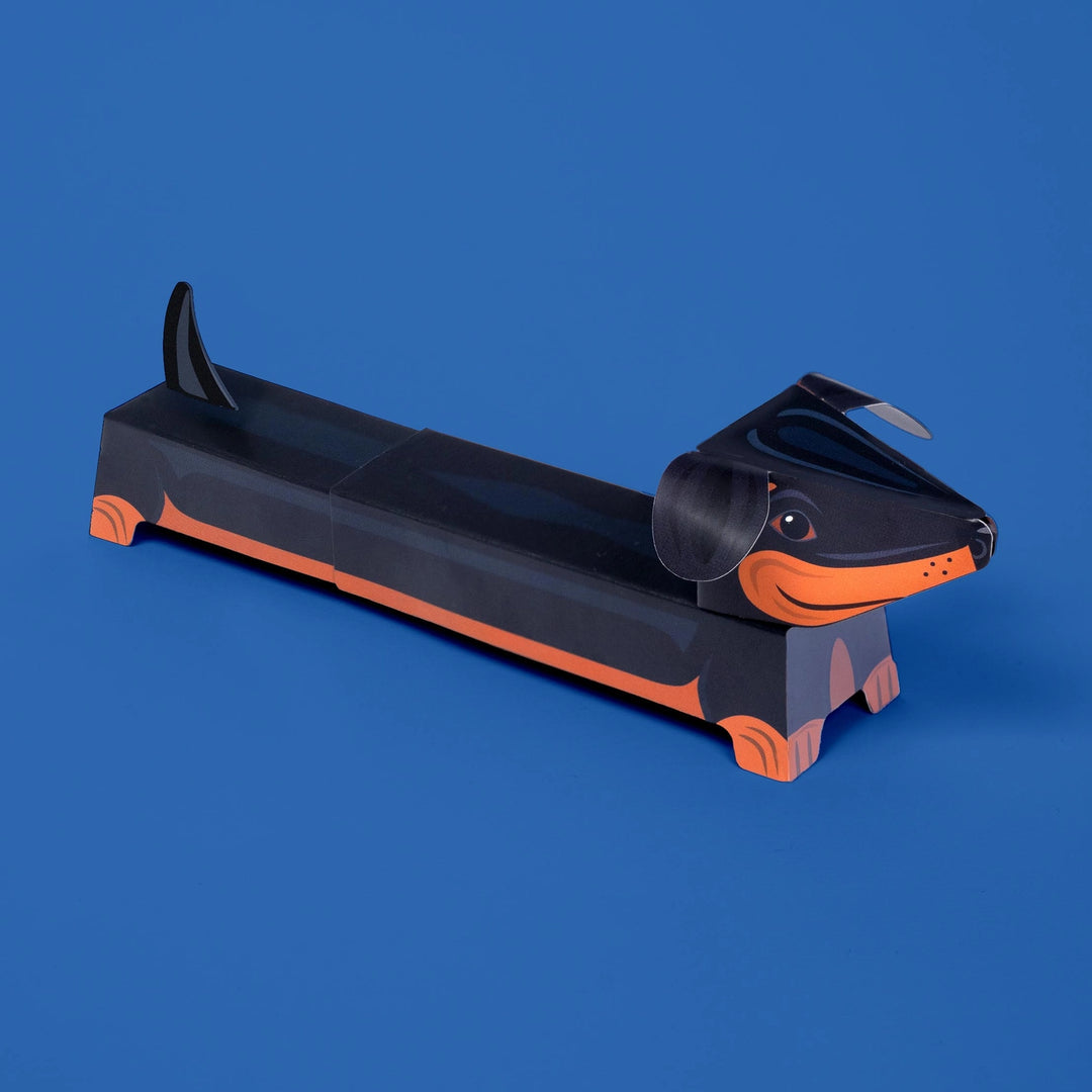 A dachshund-shaped black and orange paper model photographed on a solid blue background.
