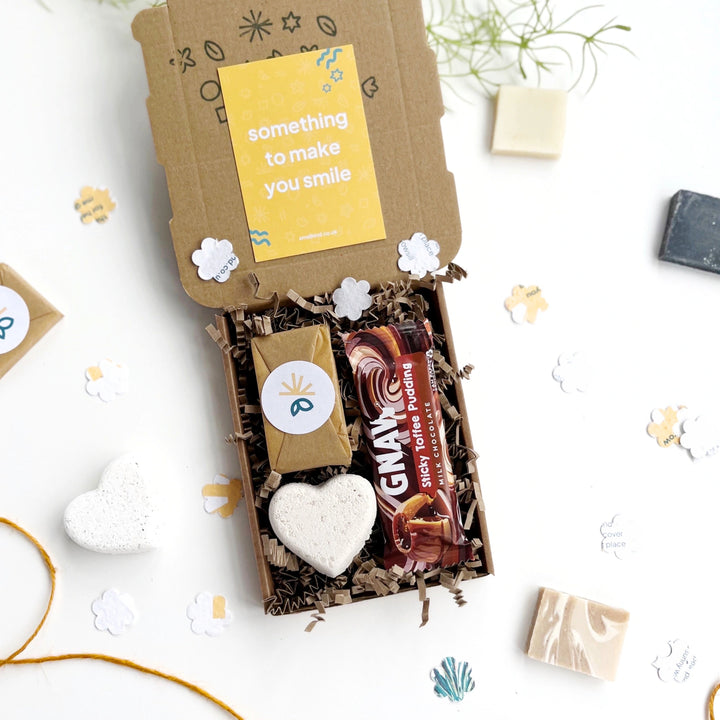Deluxe Hug in a Box Gift with chocolate, heart shaped bath bomb, wrapped mini soap bar nestled in shredded paper. There are paper flowers scattered and and a card which reads 'something to make you smile'